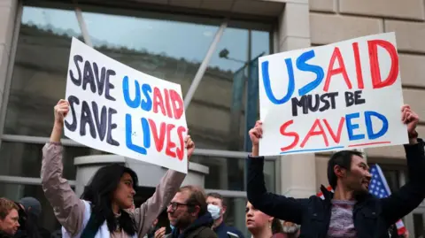 Getty Images People stand in front of the US Agency for International Development (USAID) with placards saying "Save USAID, Save Lives"