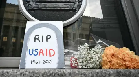 AFP A sign placed reading "RIP USAID 1961-2925" and flowers are placed beneath the covered seal of the US Agency for International Development headquarters in Washington, DC