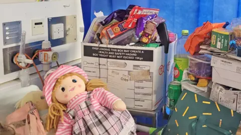 Two cloth dolls, one with blond woollen hair and a stripy pink hat, lie on Jessie's hospital bed next to the usual hospital tubes and oxygen outlets. On the bedside table are drinks and cardboard boxes piled high with biscuits, snacks, art materials and other possessions.