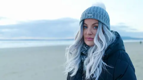 Joanne, who has fashionably dyed grey hair and blue eyes, and wears a grey woolly hat and black coat, looks seriously at the camera on an empty Welsh beach at dusk