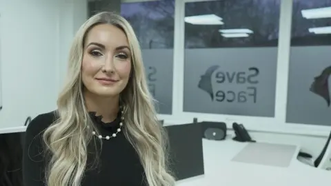 Ashton Collins, long blonde hair, a black top, white necklace, sits in her white office. Visible on the window is the logo of her organisation, Save Face.