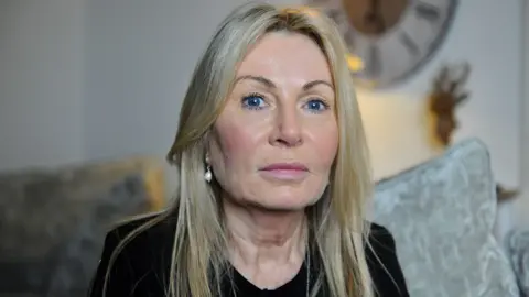 Janet Taylor, with long blonde hair and blue eyes, wearing pearl earrings, sitting on the grey couch in her living room, looking straight at the camera