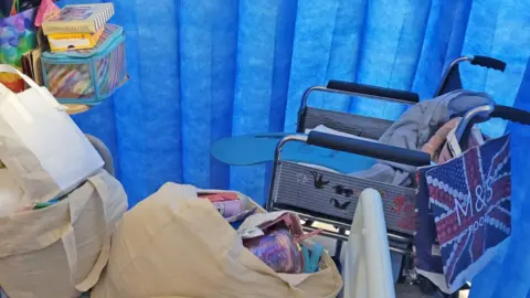 Jessie's wheelchair, decorated with stickers, is parked next to the hospital bed, inside the drawn blue curtain. On her bed are some canvas tote bags overflowing with her colourful possessions.