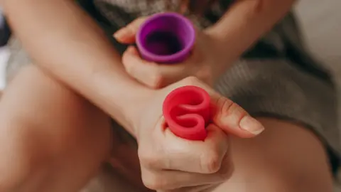 Getty Images A girl with two menstrual cups in either hand, one purple and one red. The hand in the foreground is squeezing the flexible red cup into a C shape.